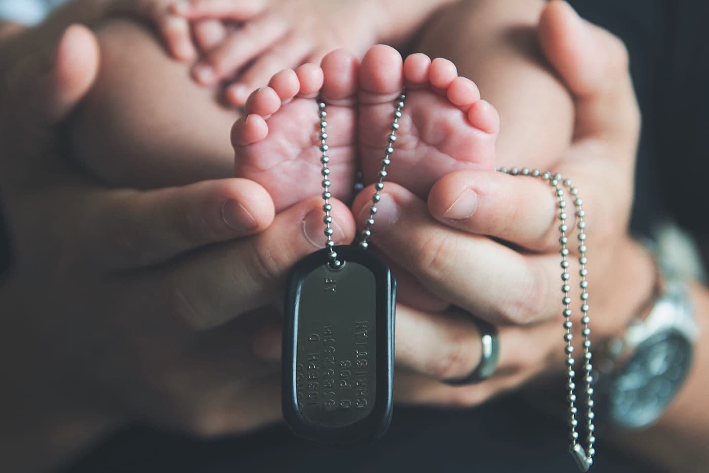 A parent cradles their baby's feet, which are draped with military tags.